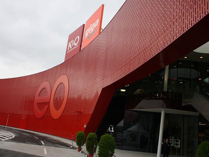 Ein modernes Gebäude mit einer großen, geschwungenen roten Fassade mit den Buchstaben „eo“. Auf dem Dach sind Schilder für K&O und Giga Sport zu sehen. Der Eingang ist aus Glas, daneben stehen Topfpflanzen, und im Vordergrund verläuft eine Kurve.