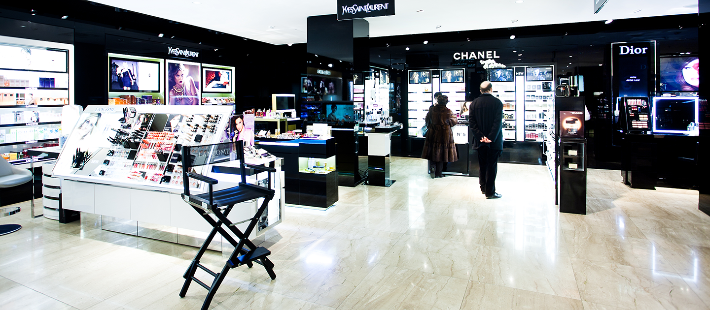 Ein hell erleuchteter Kosmetikladen mit Auslagen für Yves Saint Laurent, Chanel und Dior. Käufer stöbern in den Produkten auf eleganten Regalen und Theken. Im Vordergrund ist der Stuhl eines Maskenbildners zu sehen. Der Boden ist glänzend und sauber.