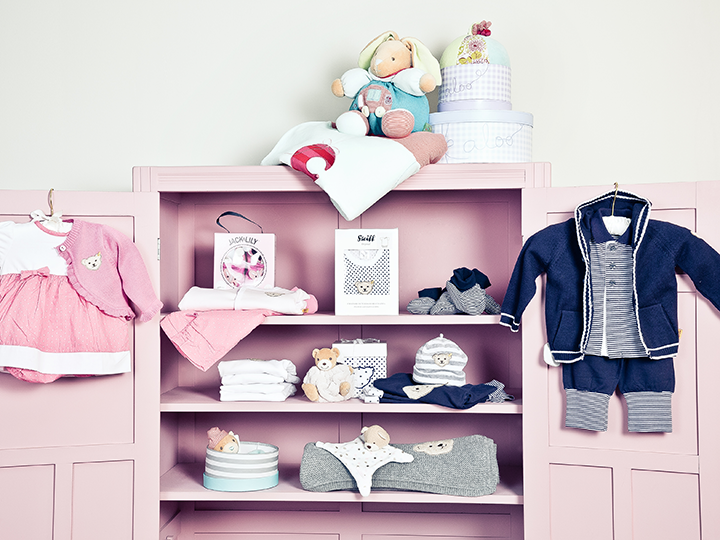 Ein pastellrosa Schrank mit Babykleidung und Accessoires, darunter ein rosa Kleid, ein blaues Outfit, Stofftiere, Babyschuhe, Mützen und eine Decke. Ein paar Stofftiere und Geschenkboxen stehen oben auf dem Schrank.