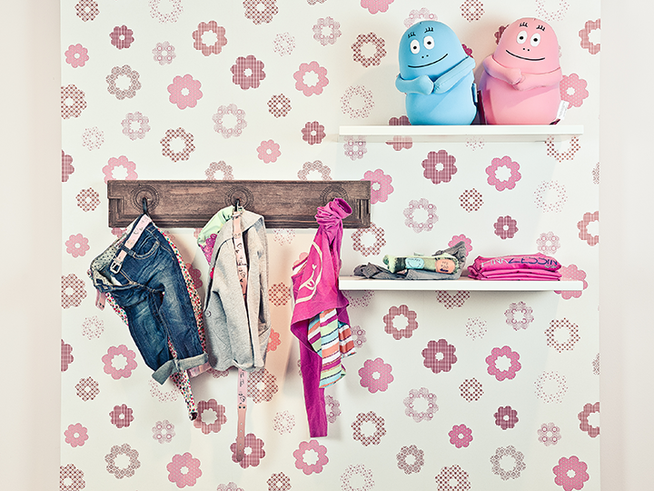Ein Kinderzimmer mit geblümter Tapete und einem Holzregal, an dem kleine Kleidungsstücke an Haken hängen. Darüber befinden sich auf zwei Regalen gefaltete Kleidungsstücke und zwei blau-rosa Cartoon-Behälter.