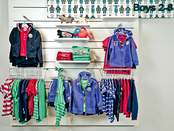 Ein Schaufenster in einem Kinderbekleidungsgeschäft mit verschiedenen Kapuzenpullovern und Hemden in Blau, Rot, Grün und mit Streifenmustern. Einige hängen auf Kleiderbügeln, andere liegen gefaltet auf weißen Regalen. Kleine Spielzeuge schmücken das oberste Regal.