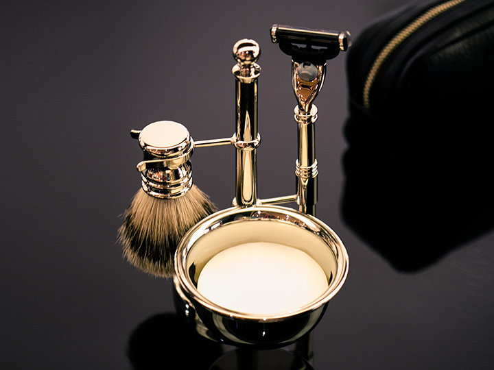 Ein luxuriöses Rasierset aus Gold umfasst einen Pinsel, einen Ständer mit Rasierer und eine Seifenschale, alles auf einer glänzenden schwarzen Oberfläche.