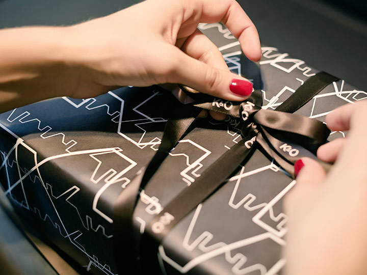 Hände mit rotem Nagellack binden eine schwarze Schleife an ein in dunkles Papier mit abstraktem weißen Linienmuster eingewickeltes Geschenk.