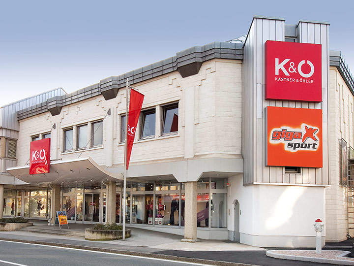 Das moderne Gebäude mit beiger Fassade beherbergt die Geschäfte K&Ö Kastner & Öhler und Giga X Sport. Rote und orangefarbene Schilder sind auffällig. Große Fenster und eine Straße davor unter klarem Himmel runden das Bild ab.
