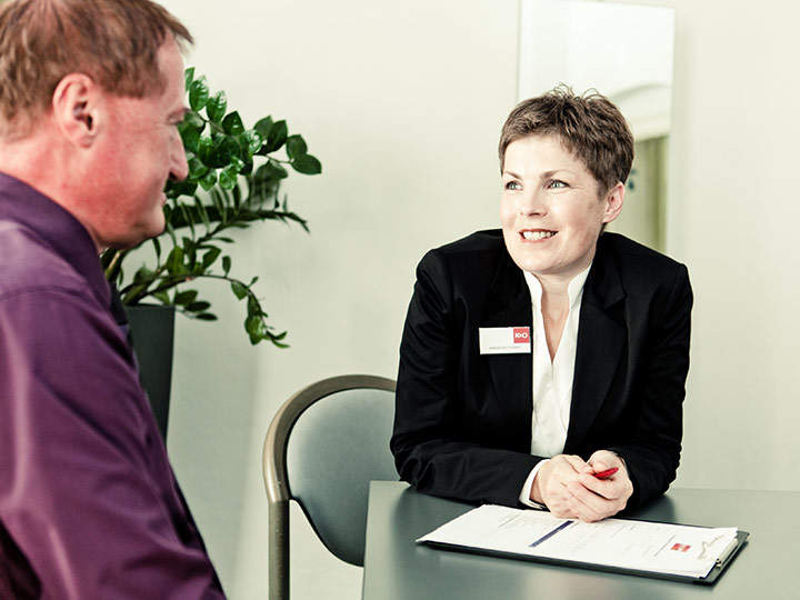 Eine Frau in einem schwarzen Blazer und einem weißen Hemd sitzt lächelnd mit einem Mann in einem lila Hemd an einem Schreibtisch. Sie unterhalten sich. Im Hintergrund steht neben einer weißen Wand eine Topfpflanze.