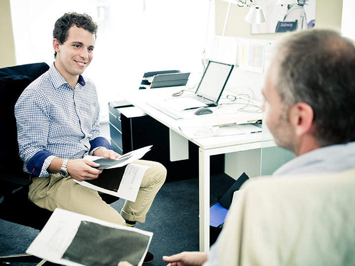 Zwei Männer sitzen an einem Schreibtisch in einem Büro. Ein Mann hält Papiere in der Hand und lächelt, während der andere zuhört. Im Hintergrund stehen Computer und Büromaterial, was eine professionelle Atmosphäre schafft.