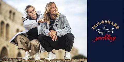 Ein Mann und eine Frau hocken im Freien auf einer felsigen Oberfläche. Beide tragen legere Yachtkleidung mit leichten Jacken. Die Frau zeigt in die Ferne. Im Hintergrund ist ein altes Steingebäude zu sehen und rechts das Logo von Paul & Shark Yachting.