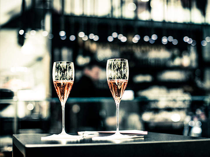 Zwei Gläser Champagner auf einem Tisch in einer schwach beleuchteten Bar. Der Hintergrund ist verschwommen, mit dekorativen Lichtern und Regalen, die eine gemütliche Atmosphäre schaffen.