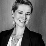 Eine lächelnde Person mit kurzen Haaren, verzierten Ohrringen und einem eleganten schwarzen Blazer über einer weißen Bluse steht vor einem strukturierten Hintergrund. Das Bild ist in Schwarzweiß.