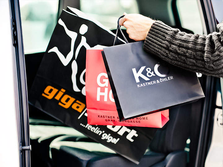 Eine Person im Pullover legt Einkaufstüten mit den Logos verschiedener Marken, darunter Kastner & Öhler und Giga Sport, auf den Rücksitz eines Fahrzeugs. Die Tüten sind schwarz und rot, was auf einen kürzlichen Einkaufsbummel schließen lässt.