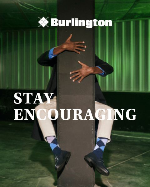 Eine Person in einem dunklen Anzug und blauen Argyle-Socken umarmt spielerisch eine Säule in einer industriellen Umgebung. Die Worte „Stay Encouraging“ sind darüber gelegt, mit „Burlington“ oben. Der Hintergrund ist grün beleuchtet.
