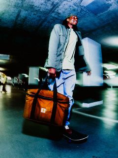 Mann, der in einem Parkhaus Skateboard fährt, eine große braune Carhartt-Tasche trägt, eine grüne Jacke und Jeans bei künstlichem Licht trägt.