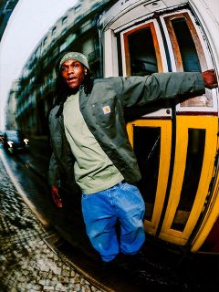 Ein Mann in legerer Kleidung lehnt sich auf einer Kopfsteinpflasterstraße aus einer gelben Straßenbahn, wobei die Gebäude der Stadt im Hintergrund verschwimmen.