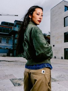 Eine Frau mit langen dunklen Haaren, grüner Steppjacke und hellbrauner Hose steht in einer städtischen Umgebung im Freien und schaut über ihre Schulter.