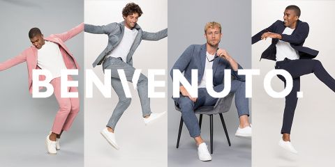 Vier Männer in eleganten Anzügen und weißen Turnschuhen sind vor einem hellen Hintergrund zu sehen. Zwei springen energisch, einer lümmelt auf einem Stuhl und einer posiert. Das Wort „BENVENUTO“ ist über das Bild gelegt.