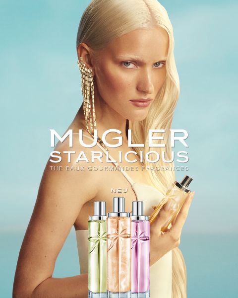 Eine Frau mit langen, platinblonden Haaren hält ein Parfümfläschchen in der Hand. Vor ihr sind drei farbenfrohe „Mugler Starlicious“-Parfüms ausgestellt.