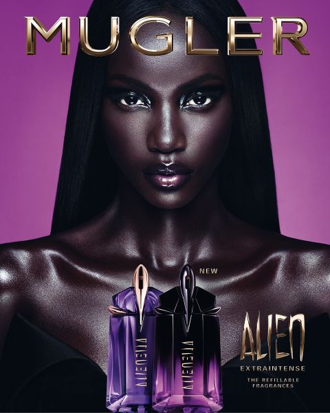 Eine Frau mit strahlend dunkler Haut und glattem schwarzen Haar posiert vor einem violetten Hintergrund. Vor ihr stehen zwei Alien-Parfümflakons von Mugler. Der Text lautet „MUGLER“ und „ALIEN EXTRA INTENSE THE REFILLABLE FRAGRANCES“.