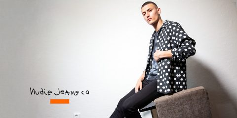Eine Person sitzt auf einem Stuhl, trägt ein schwarz-weiß gepunktetes Hemd und schwarze Jeans und blickt in die Kamera. Auf der linken Seite des Bildes erscheint der Text „Nudie Jeans Co“. Die Szene ist in einem Innenbereich vor einer schlichten Wand.