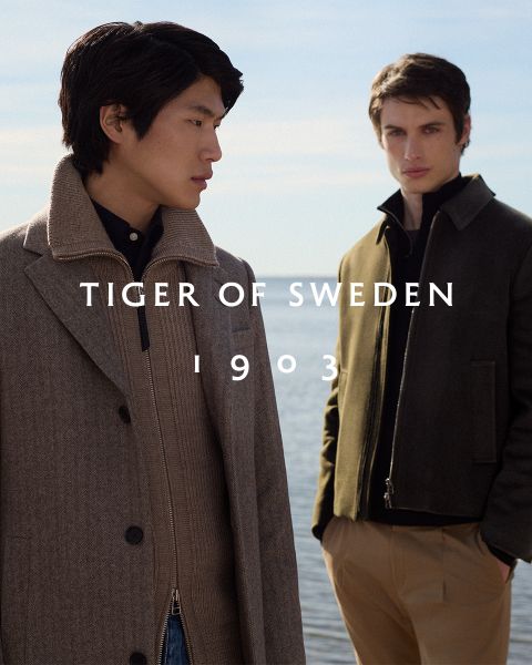 Zwei Männer in eleganten Mänteln stehen im Freien am Wasser unter einem klaren Himmel. Der Text "TIGER OF SWEDEN 1903" steht mittig auf dem Bild.