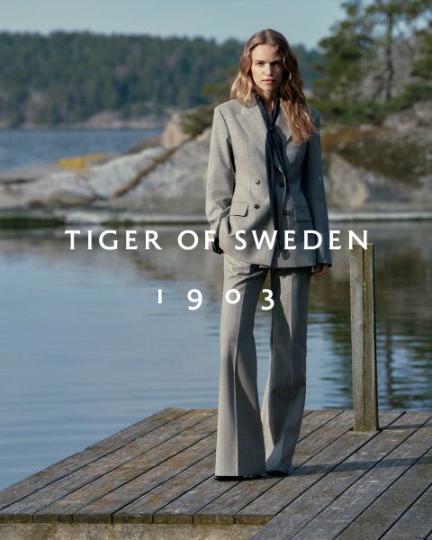 Eine Frau in einem grauen Anzug steht auf einem hölzernen Steg an einem See mit felsigen, baumbestandenen Ufern. Der Text "TIGER OF SWEDEN 1903" ist quer über das Bild geschrieben.