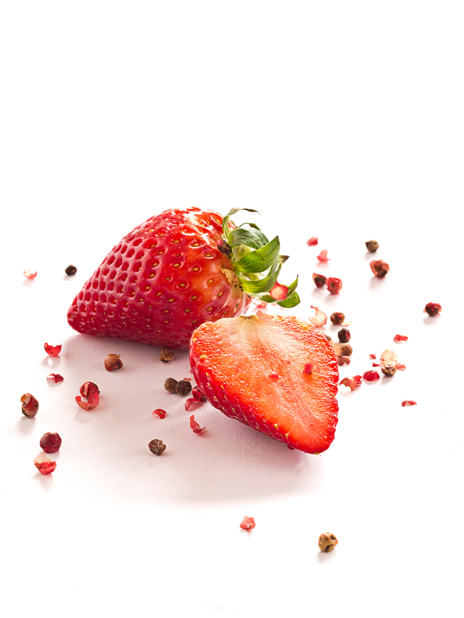 Eine reife Erdbeere wird halbiert und auf eine weiße Fläche gelegt, umgeben von verstreuten rosa Pfefferkörnern. Die Kerne und die leuchtend rote Farbe der Erdbeere sind deutlich zu erkennen.