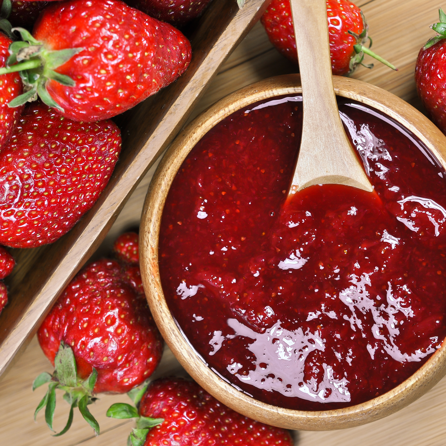 Eine Holzschale gefüllt mit leuchtend roter Erdbeermarmelade, in der ein Löffel liegt. Frische Erdbeeren sind in der Schale verstreut und zeigen ihre leuchtend rote Farbe und ihre grünen Blätter, alles auf einer Holzoberfläche.