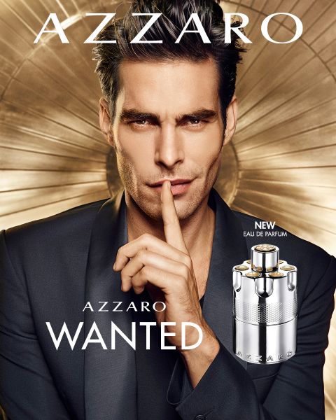 Ein Mann im Anzug legt seinen Finger in einer beschwichtigenden Geste an die Lippen. Vor ihm steht eine Flasche Azzaro Wanted Eau de Parfum. Der Hintergrund zeigt ein metallisches, kreisförmiges Design. Der Text lautet „Azzaro“ und „Wanted“.
