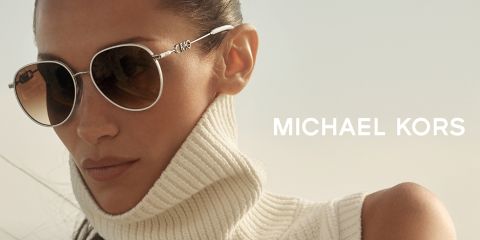 Eine Person mit runder Sonnenbrille und weißem Rollkragenpullover blickt zur Seite. Der Hintergrund ist weich verschwommen und rechts ist in weißer Schrift der Name „Michael Kors“ zu sehen.