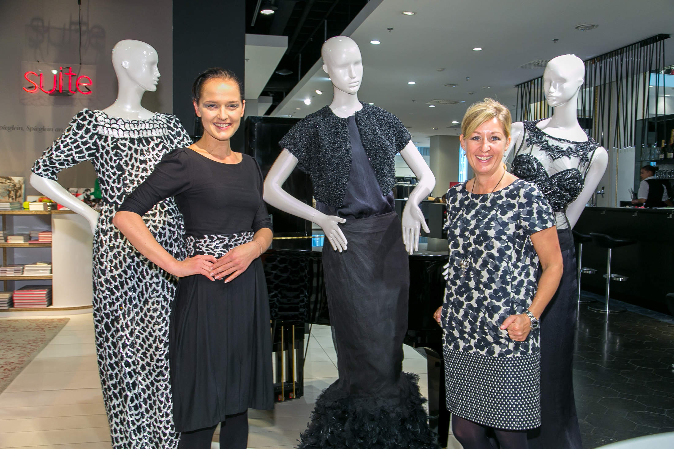 Zwei Frauen in gemusterten schwarz-weißen Outfits stehen lächelnd neben Schaufensterpuppen in eleganten schwarzen Ensembles in einem schicken Boutique-Ambiente.