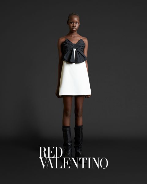 Eine Person posiert vor einem dunkelgrauen Hintergrund und trägt ein schwarz-weißes Outfit mit einer großen Schleife oben drauf. Sie trägt schwarze kniehohe Stiefel. Unten ist der Text „RED VALENTINO“ zu sehen.