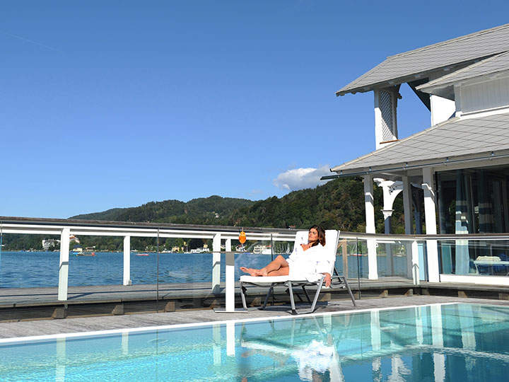 Eine Person liegt auf einem Stuhl neben einem Pool und genießt einen Drink unter einem klaren blauen Himmel. Die Umgebung bietet Ausblick auf einen ruhigen See mit grünen Hügeln und einem teilweise sichtbaren Gebäude in der Nähe.