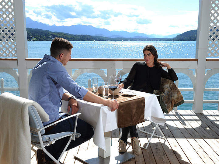 Ein Mann und eine Frau sitzen sich an einem Tisch auf einer Holzterrasse an einem See gegenüber. Die Sonne scheint hell und im Hintergrund sind Berge zu sehen. Beide haben Getränke auf dem Tisch und die Frau trägt einen Pelzmantel.