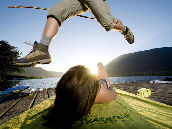 Eine Person springt über eine andere Person, die auf einem Holzsteg an einem See liegt. Im Hintergrund sind Berge und die Sonne untergehend zu sehen. Die Person auf dem Steg liegt auf einem grünen Handtuch und in der Nähe liegen Badesachen.