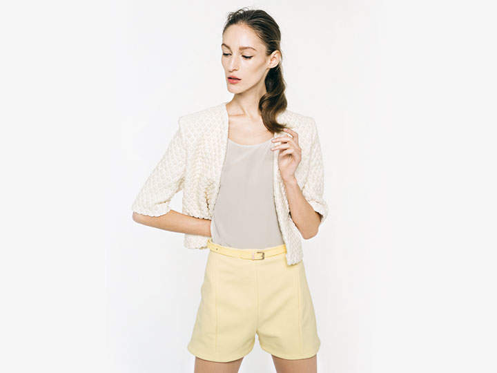 Eine Person steht vor einem hellen Hintergrund und trägt einen strukturierten weißen Blazer, ein beiges Oberteil und hellgelbe Shorts. Sie hat langes Haar, das zur Seite frisiert ist, und blickt mit heiterem Gesichtsausdruck nach unten.