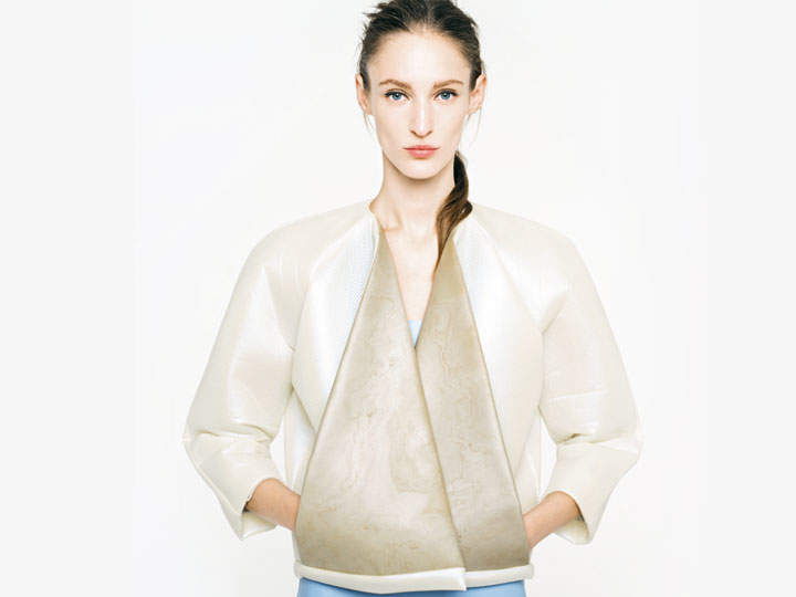 Eine Person mit zurückgekämmten langen Haaren trägt eine stilvolle, strukturierte Jacke in Creme und Beige. Die Jacke hat breite Revers und ein minimalistisches Design. Der Hintergrund ist schlicht weiß und unterstreicht den modernen Modelook.