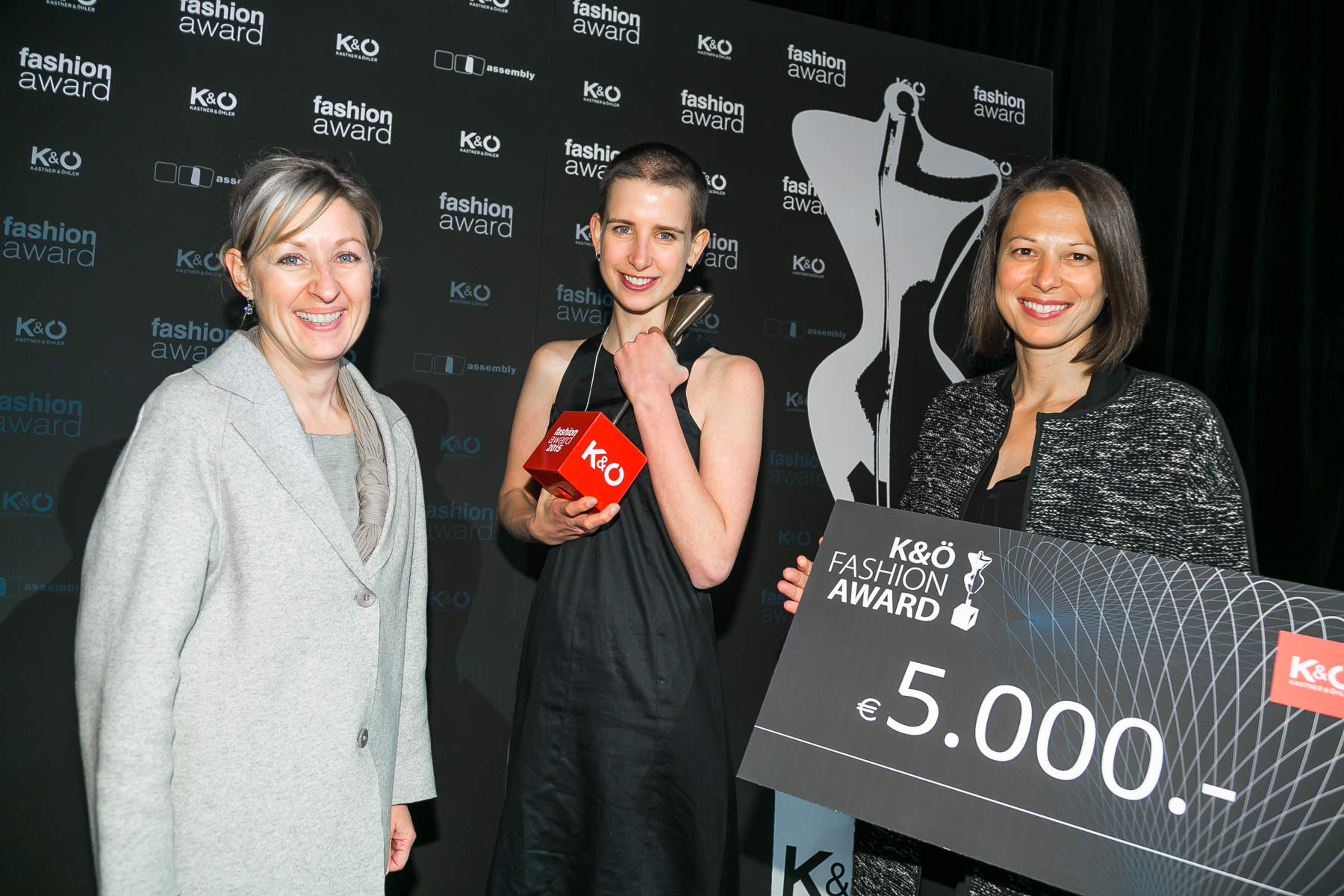 Drei lächelnde Frauen bei einer Modepreisverleihung. Eine hält eine Trophäe, eine andere einen großen Scheck über 5.000 Euro. Sie stehen vor einem Hintergrund mit dem Text „K&Ö Fashion Award“.