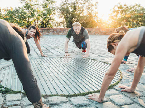 Eine Gruppe von Menschen macht im Freien auf einem gepflasterten Platz im Kreis Liegestütze. Sie lächeln und sind von Grün umgeben, im Hintergrund scheint die Sonne.