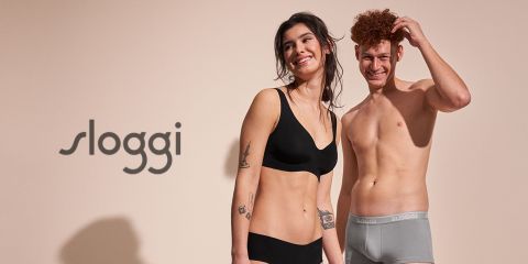 Eine lächelnde Frau und ein Mann in Unterwäsche von Sloggi stehen vor einem neutralen Hintergrund mit dem Markenlogo auf der linken Seite. Die Frau trägt ein schwarzes Set und der Mann einen grauen Slip.