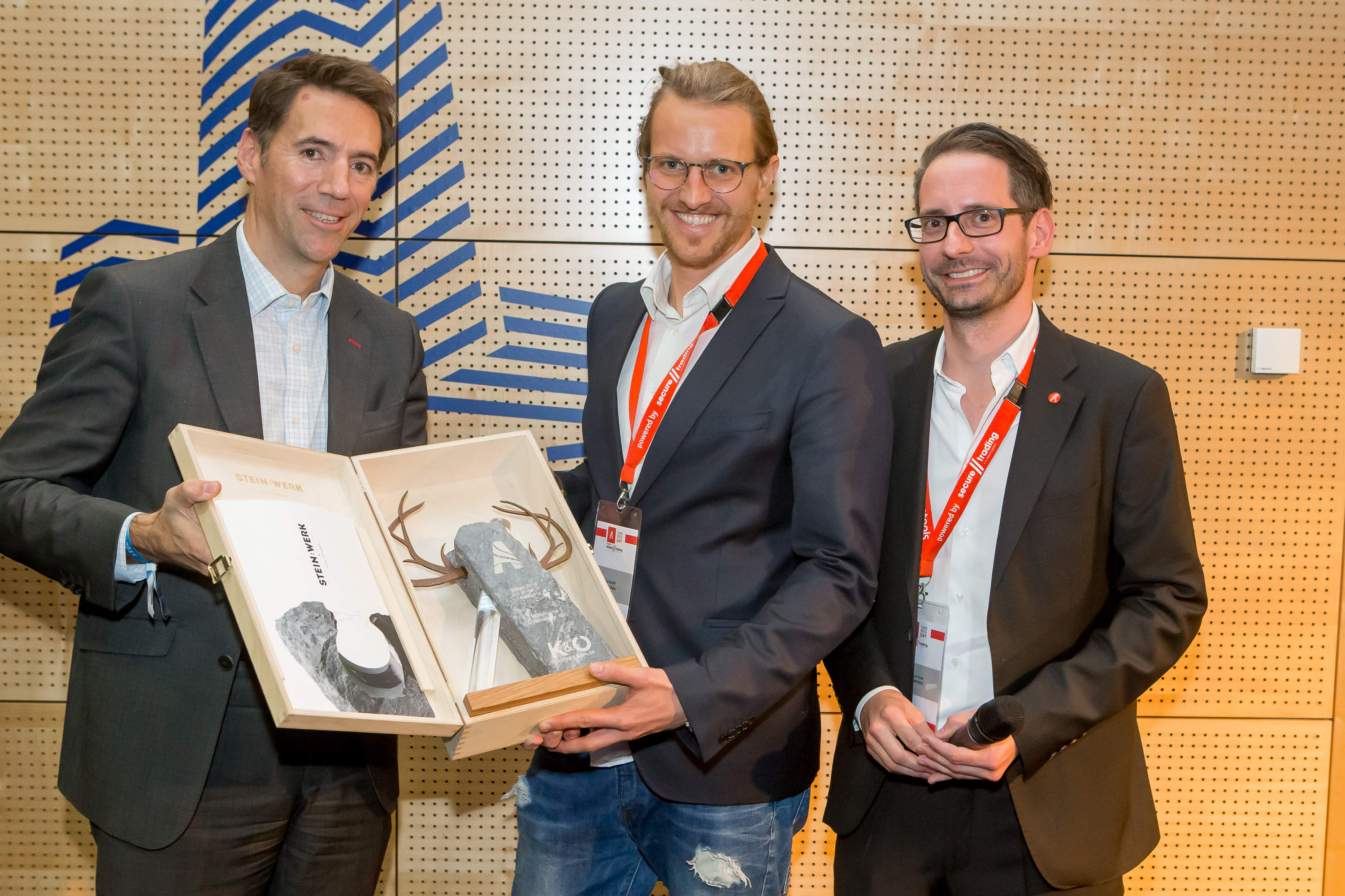 Drei Männer in Anzügen und Jeans stehen zusammen. Zwei von ihnen halten eine Geschenkbox aus Holz, die ein graues Objekt mit dem Text „KO“ und Geweihen enthält. Sie lächeln in einem Raum mit einer Holzwand und einem blauen geometrischen Logo.