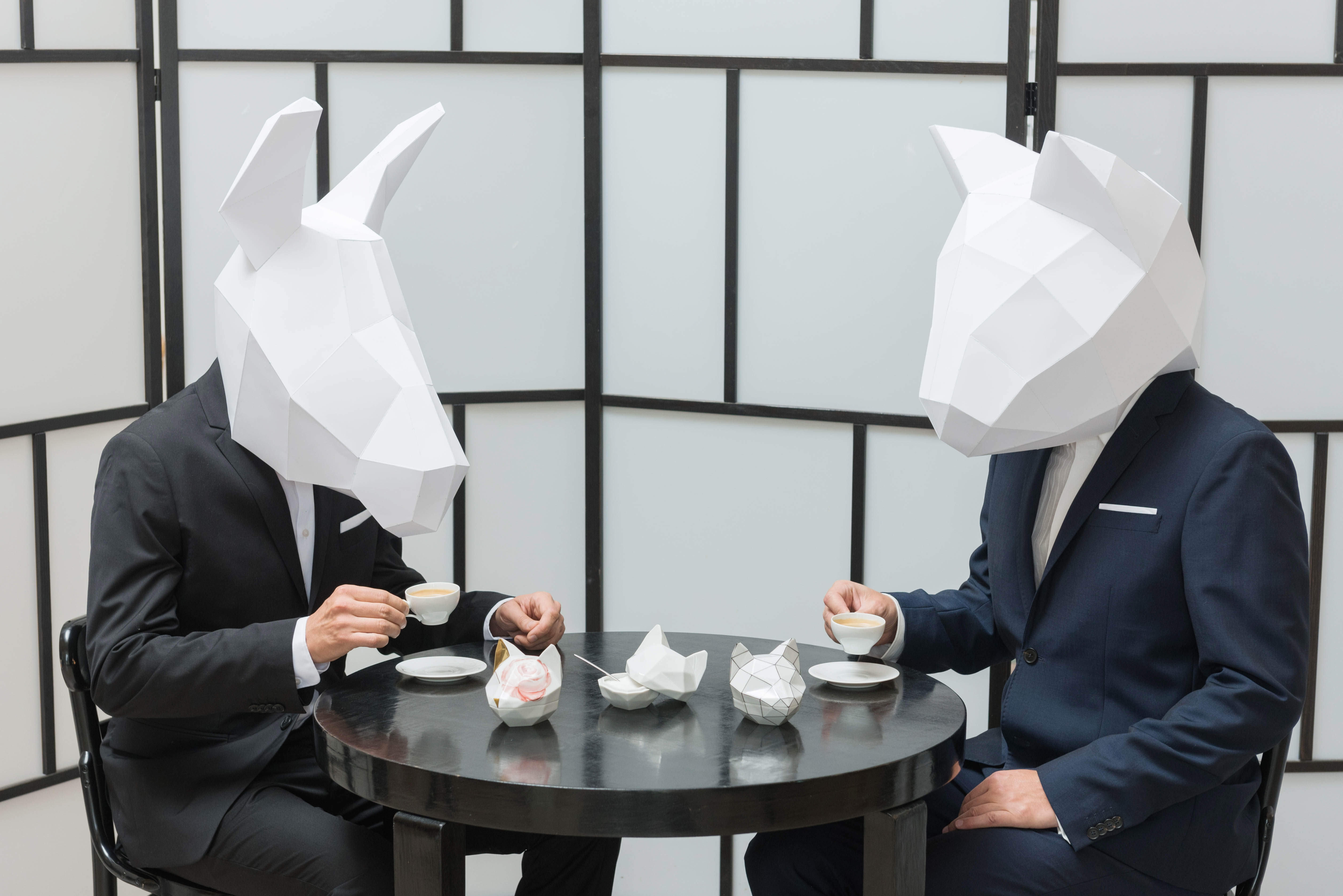Zwei Personen in Anzügen sitzen an einem runden Tisch und trinken Kaffee. Sie tragen große, geometrische Masken aus weißem Papier, die an Tierköpfe erinnern. Im Hintergrund ist ein weiß-schwarz getäfelter Paravent zu sehen. Auf dem Tisch liegen Origamistücke.