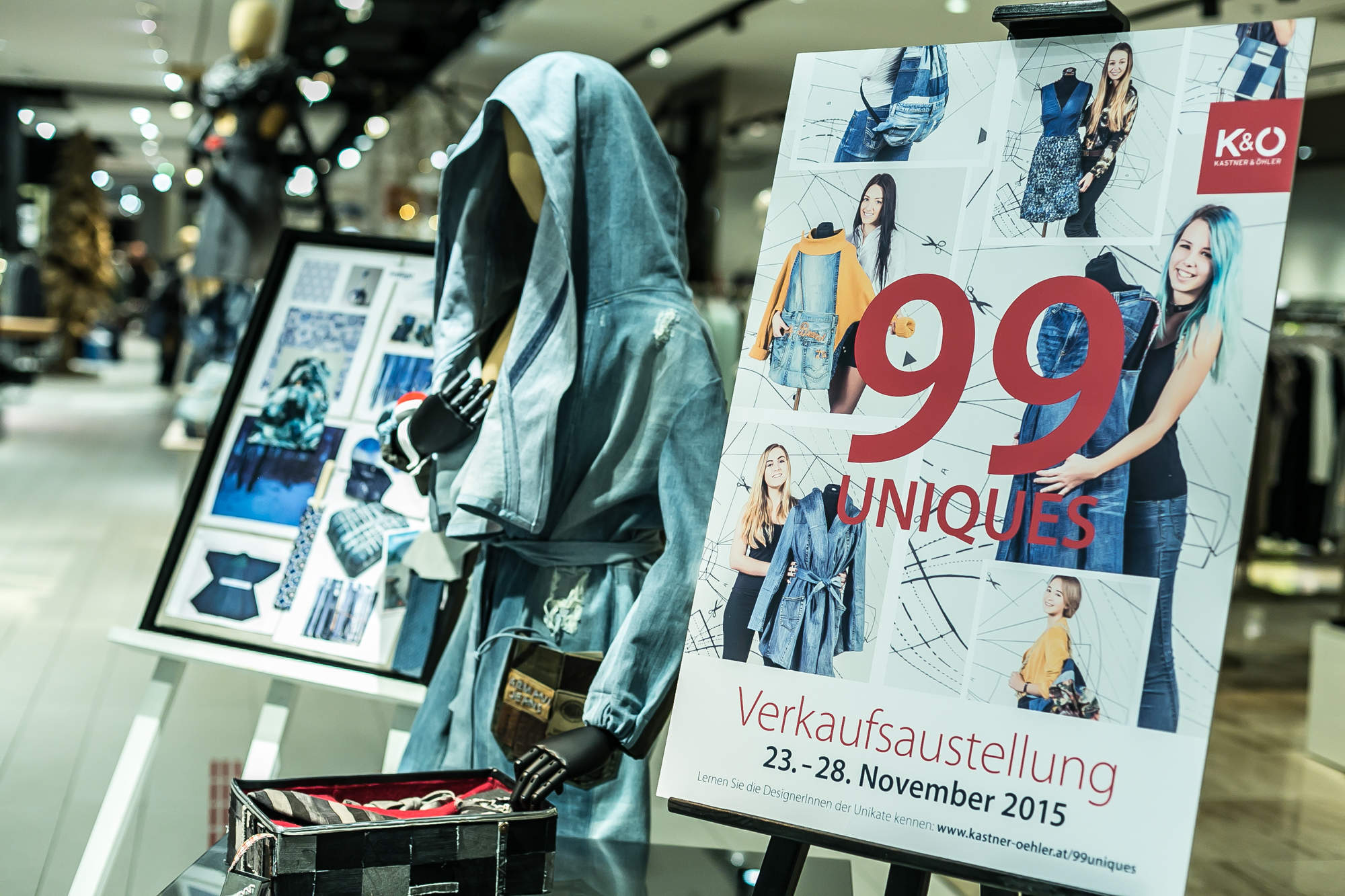 Auf einem Schaufenster sind eine Jeansjacke und ein Werbeschild für „99 Uniques“ mit Bildern verschiedener Outfits zu sehen. Die Veranstaltung findet vom 23. bis 28. November 2015 statt. Das Schaufenster befindet sich in einem hell erleuchteten Einzelhandelsumfeld.