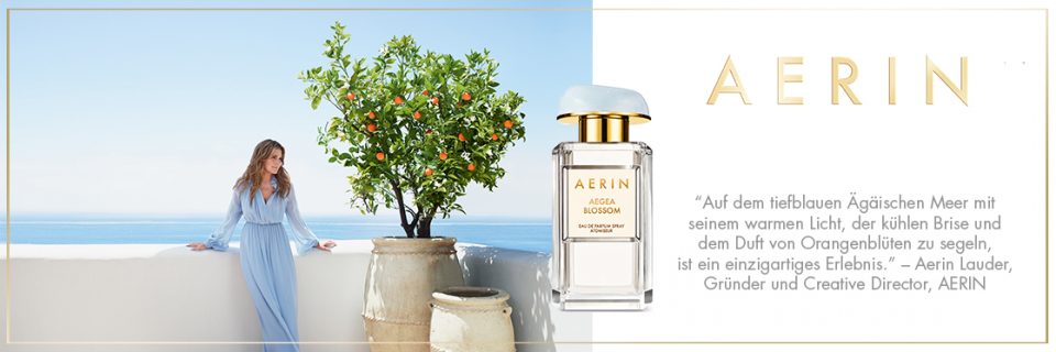 Eine Frau in einem hellblauen Kleid steht auf einer Terrasse mit Blick auf das Meer neben einem Orangenbaum. Im Vordergrund steht eine Flasche Aerin-Parfüm, auf der der Markenname deutlich zu sehen ist. Neben der Flasche ist ein deutscher Text zu sehen.
