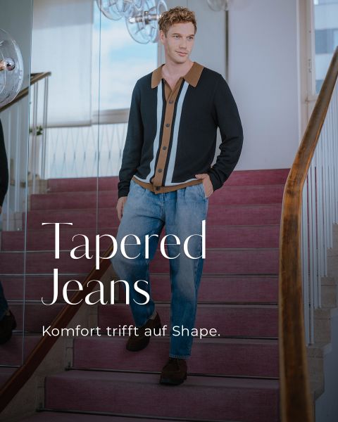 Ein Mann in einer eleganten Strickjacke und konisch zulaufenden Jeans geht eine Teppichtreppe hinunter. Der Text auf dem Bild lautet: "Tapered Jeans. Komfort trifft auf Shape." Die Umgebung ist modern mit natürlichem Licht.