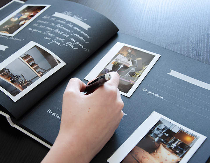 Die Hand einer Person hält einen Stift und schreibt in ein schwarzes Sammelalbum, auf dessen Seiten Fotos angebracht sind. Das Buch liegt aufgeschlagen auf einem dunklen Tisch. Die Seiten haben Platz zum Schreiben und verschiedene Bilder mit hellem Hintergrund.