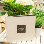 Ein weißes Fotoalbum mit den Namen „Juliane & Philipp“ auf dem Einband liegt auf einem weißen Tisch, umgeben von grünen Pflanzen und weißen Blumen.
