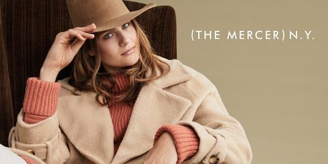 Eine Person mit beigem Mantel und Hut sitzt in einem braunen Sessel. Neben ihr steht der Text „The Mercer N.Y.“ auf einem hellbeigen Hintergrund.