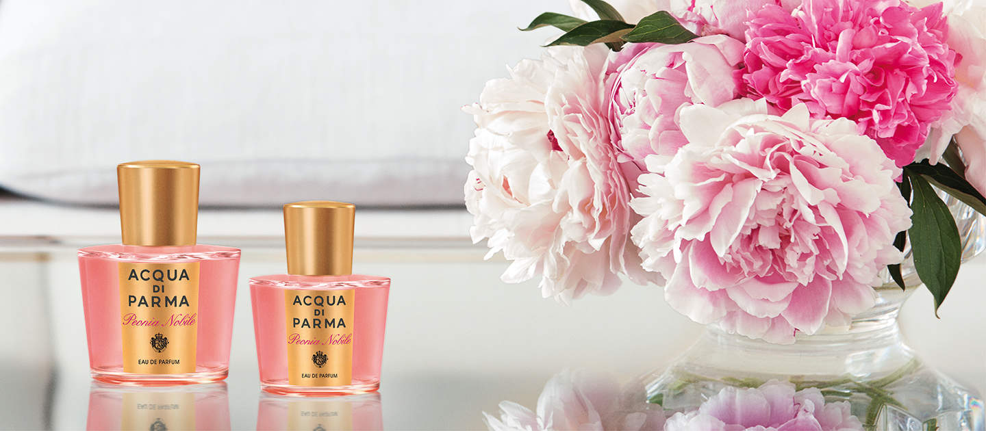 Zwei Flakons des Parfüms Acqua di Parma stehen auf einer reflektierenden Oberfläche neben einem Strauß rosa und weißer Pfingstrosen. Der Hintergrund ist unscharf und neutral gehalten, was die Eleganz und das florale Motiv hervorhebt.