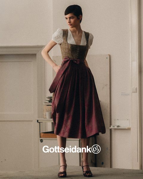 Eine Person steht drinnen und trägt ein traditionelles bayerisches Outfit, bestehend aus einer weißen Bluse, einer dekorativen Weste und einem langen lila Rock mit einer Schleife. Im Hintergrund sind eine kahle Wand und ein Stapel Bücher zu sehen. Unten ist das Wort „Gottseidank“ zu sehen.