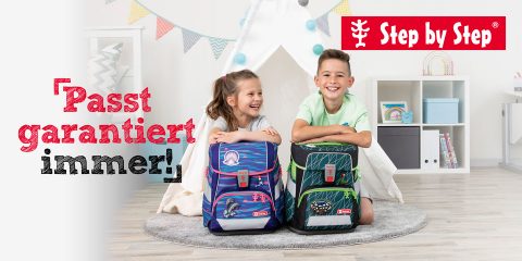 Zwei Kinder sitzen lächelnd auf einem grauen Teppich in einem Spielzimmer und haben ihre Rucksäcke vor sich. Im Hintergrund sind ein Tipi und Regale mit Spielzeug zu sehen. Der Text lautet „Passt garantiert immer!“ und „Step by Step“ mit einem roten Logo in der oberen rechten Ecke.