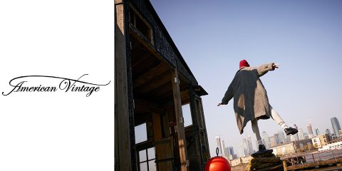 Eine Person in langem Mantel und rotem Hut balanciert auf einem Holzpfosten in der Nähe eines rustikalen Gebäudes. Im Hintergrund ist die Skyline der Stadt unter einem klaren Himmel zu sehen. Auf der linken Seite des Bildes befindet sich das Logo „American Vintage“.